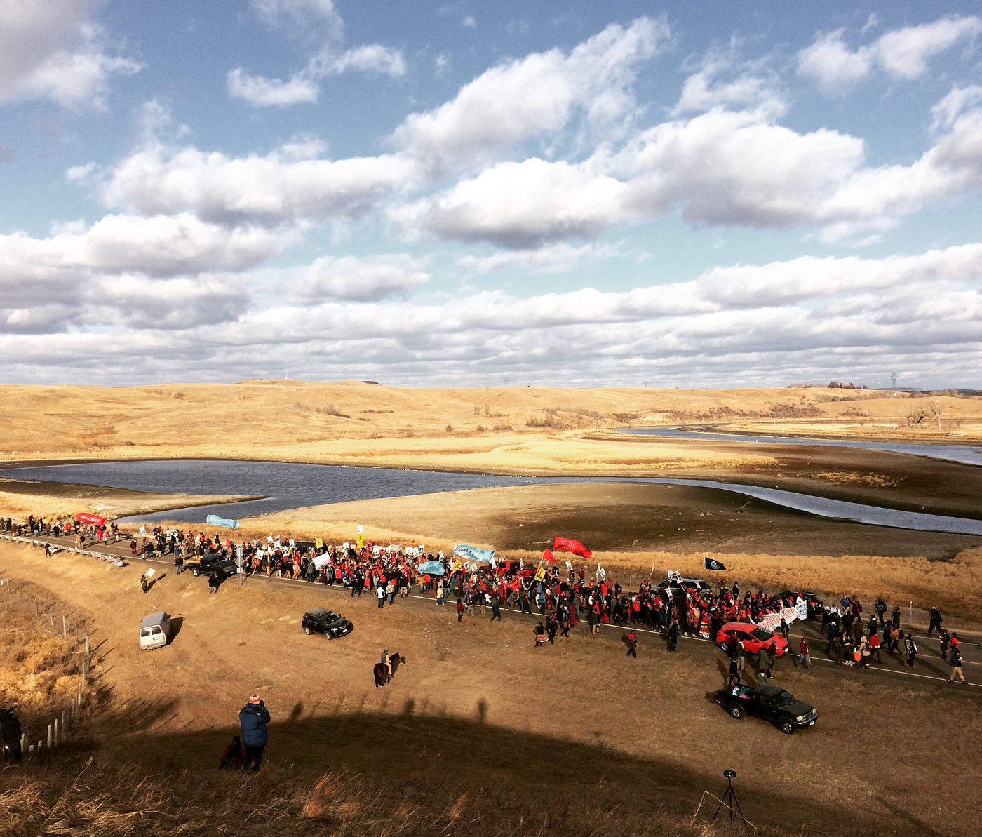 An aerial view of the encampment at Standing Rock stretched along a river valley under a wide sky.