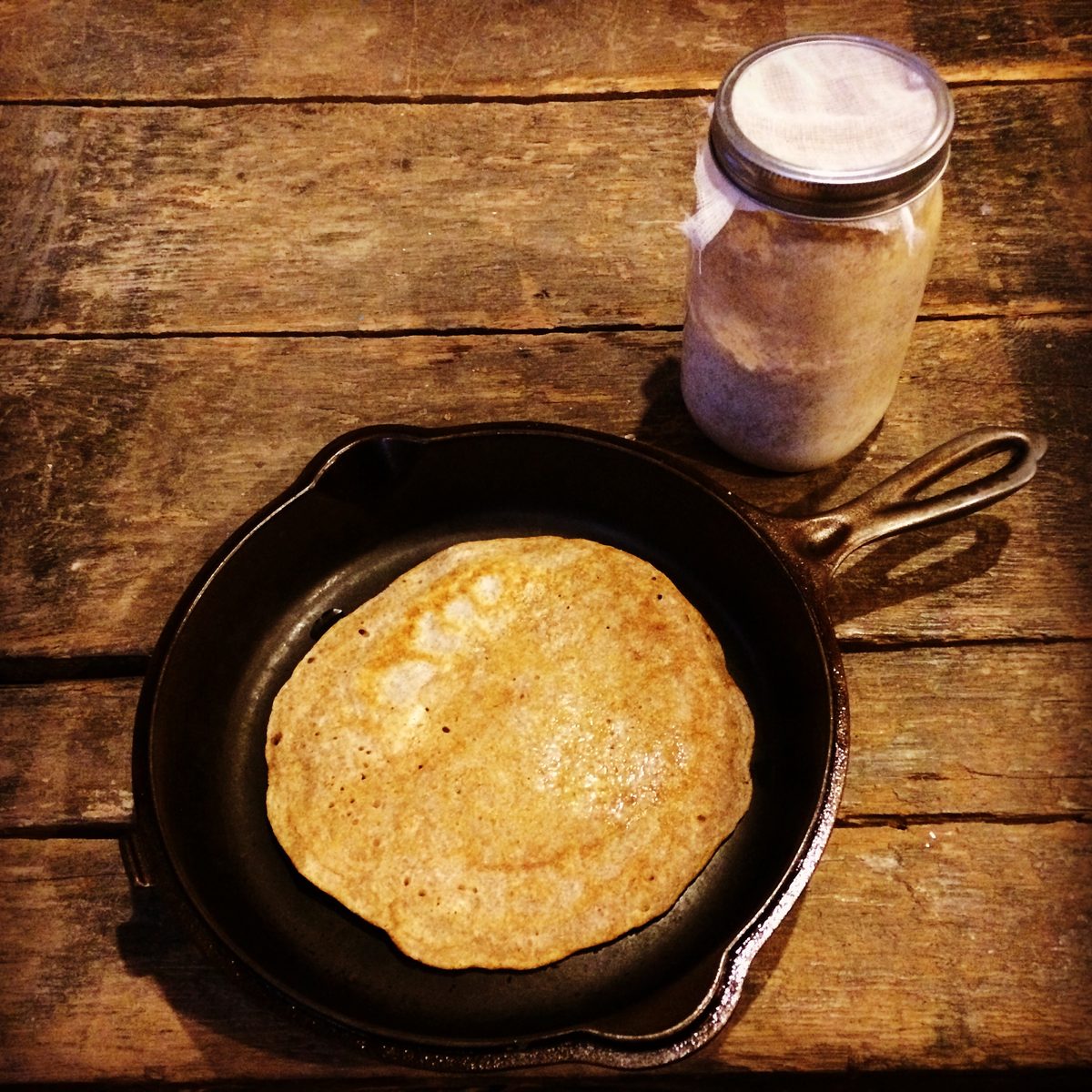 A skillet flatbread on a wooden table beside a jar of bubbling sourdough starter.