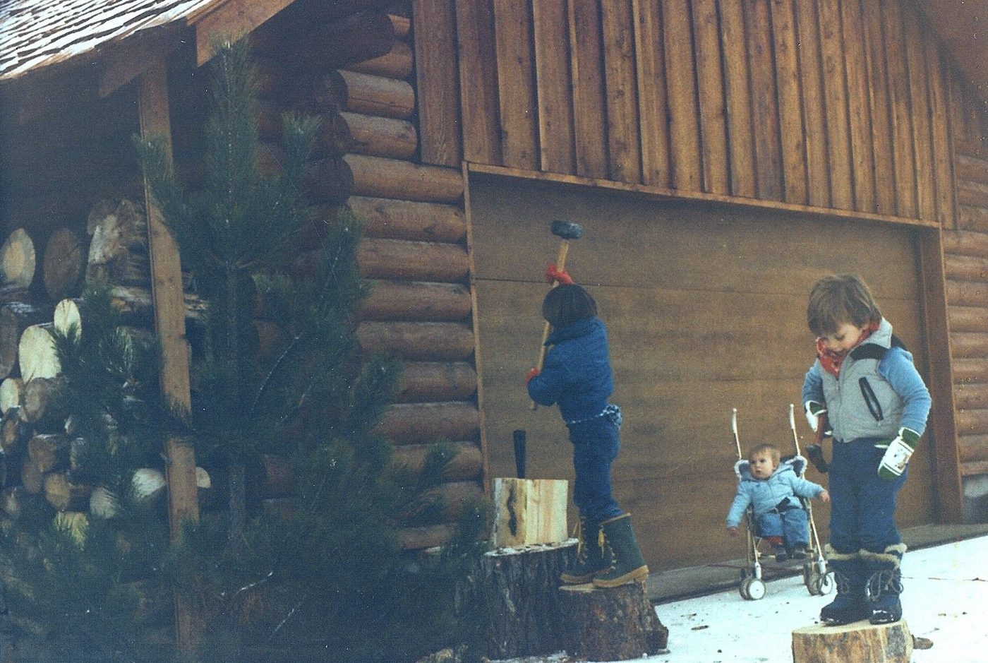 Two children in winter coats splitting wood beside a log cabin in the snow.